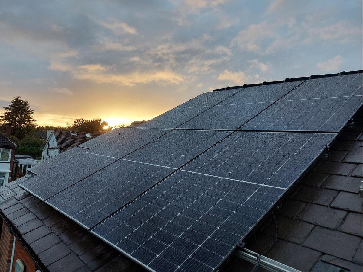 Large solar panels installed on a dark tiled roof, photographed at sunset with a cloudy sky and neighboring houses visible in the background.